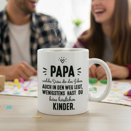 Lustige Papa-Tasse mit Spruch „wenigstens hast du keine hässlichen Kinder“, steht auf Tisch mit spielender Familie im Hintergrund