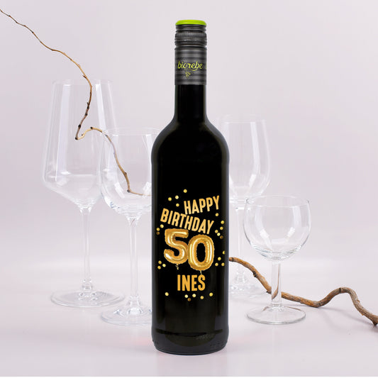 Schwarze Weinflasche mit goldener Ballonzahl „50“ und dem Text „Happy Birthday Ines“ auf dem Etikett, im Hintergrund Weingläser und Rebenzweig