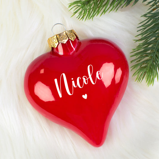 Glänzende rote Herz-Weihnachtskugel mit Name „Nicole“ auf weißem Fell, Tannenzweig im Anschnitt.