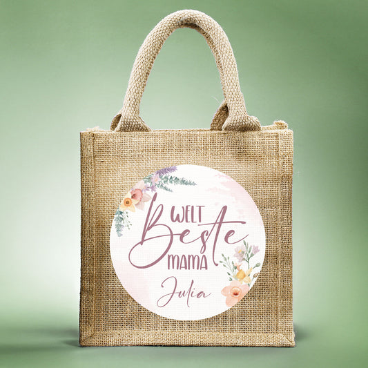Jute-Geschenktasche „Weltbeste Mama“ mit floralem Motiv & personalisierbarem Namen frontal fotografiert vor grünem Hintergrund