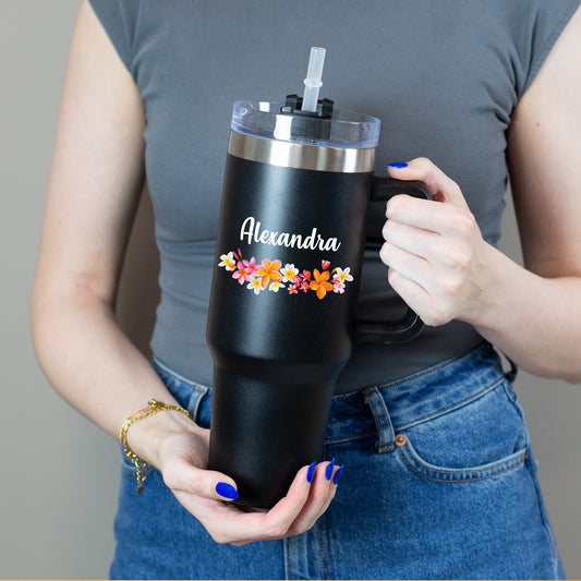Person hält Thermobecher mit buntem Blumenkranz und weißem Namen mit beiden Händen