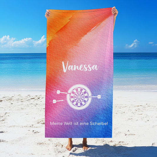 Person hält ein großes personalisiertes Badetuch mit Namen, Wunschtext und Hobby-Motiv am Strand in die Höhe – Druckmotiv gut sichtbar vor maritimer Kulisse.