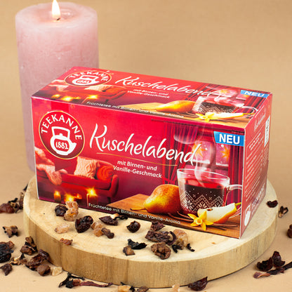 Packung „Teekanne Kuschelabend“ auf Holzscheibe platziert, dekoriert mit losen Teestückchen.