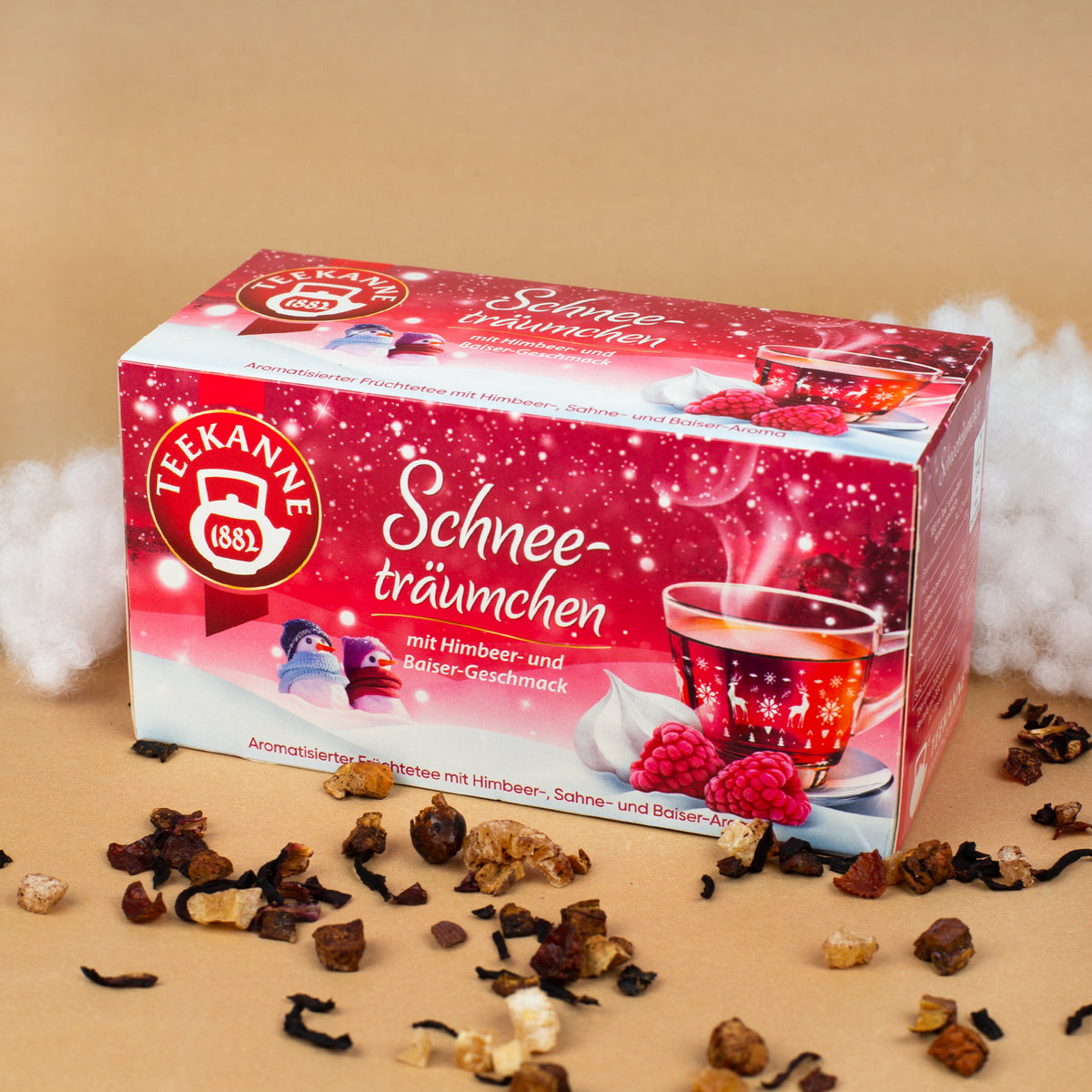 Packung „Teekanne Schneeträumchen“ dekoriert mit Watte als Schnee sowie losen Teestückchen vor hellem Hintergrund.