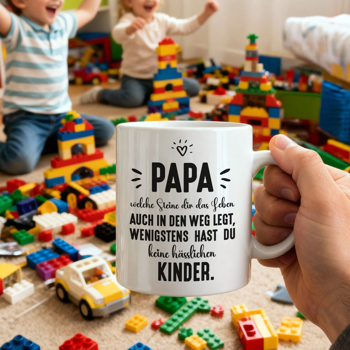 Papa-Tasse mit humorvollem Spruch, gehalten in der Hand, im Hintergrund Kinder mit bunten Bausteinen