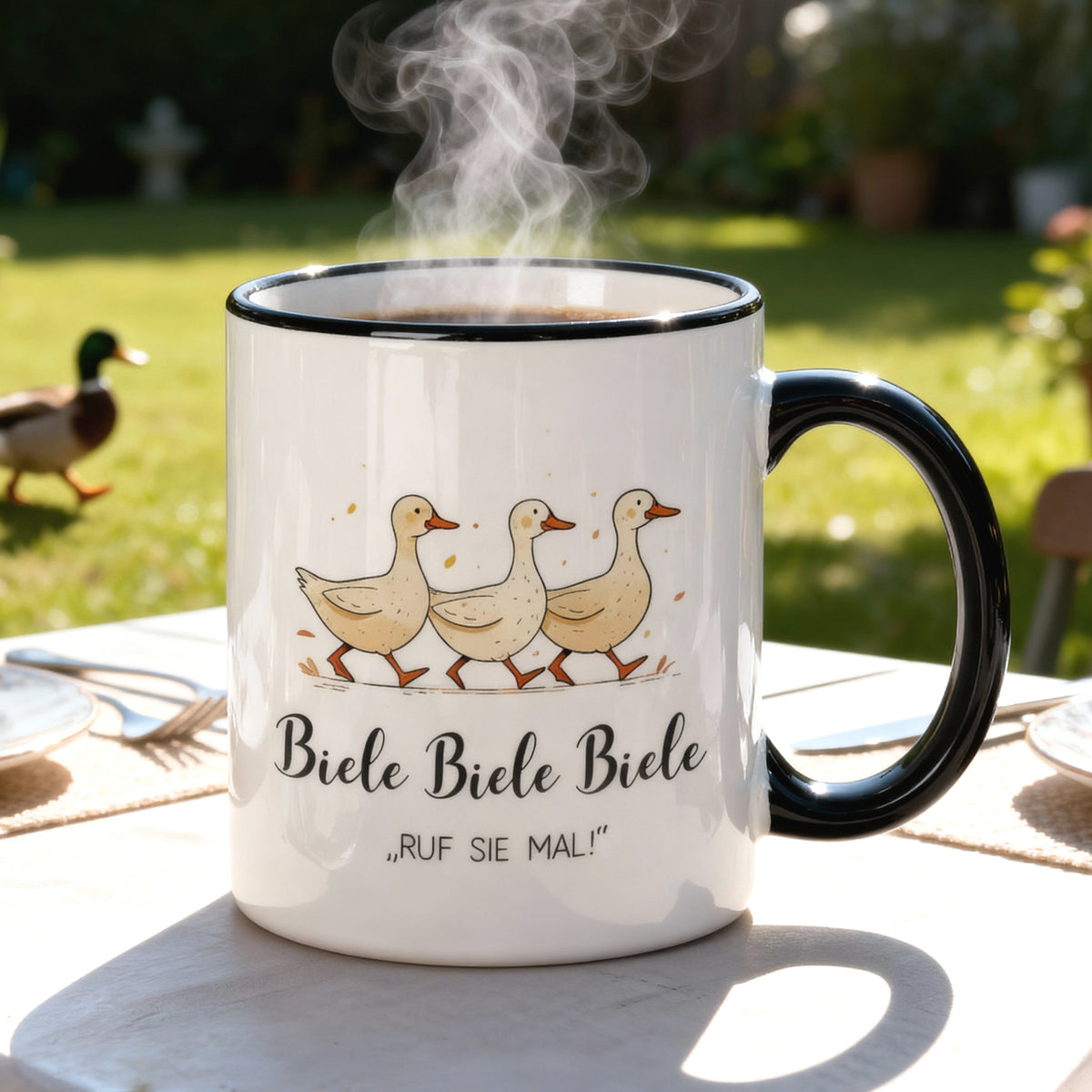 Tasse mit Entenmotiv und Spruch „Biele Biele Biele – Ruf sie mal!“ auf Gartentisch, im Hintergrund grüne Wiese mit Ente.