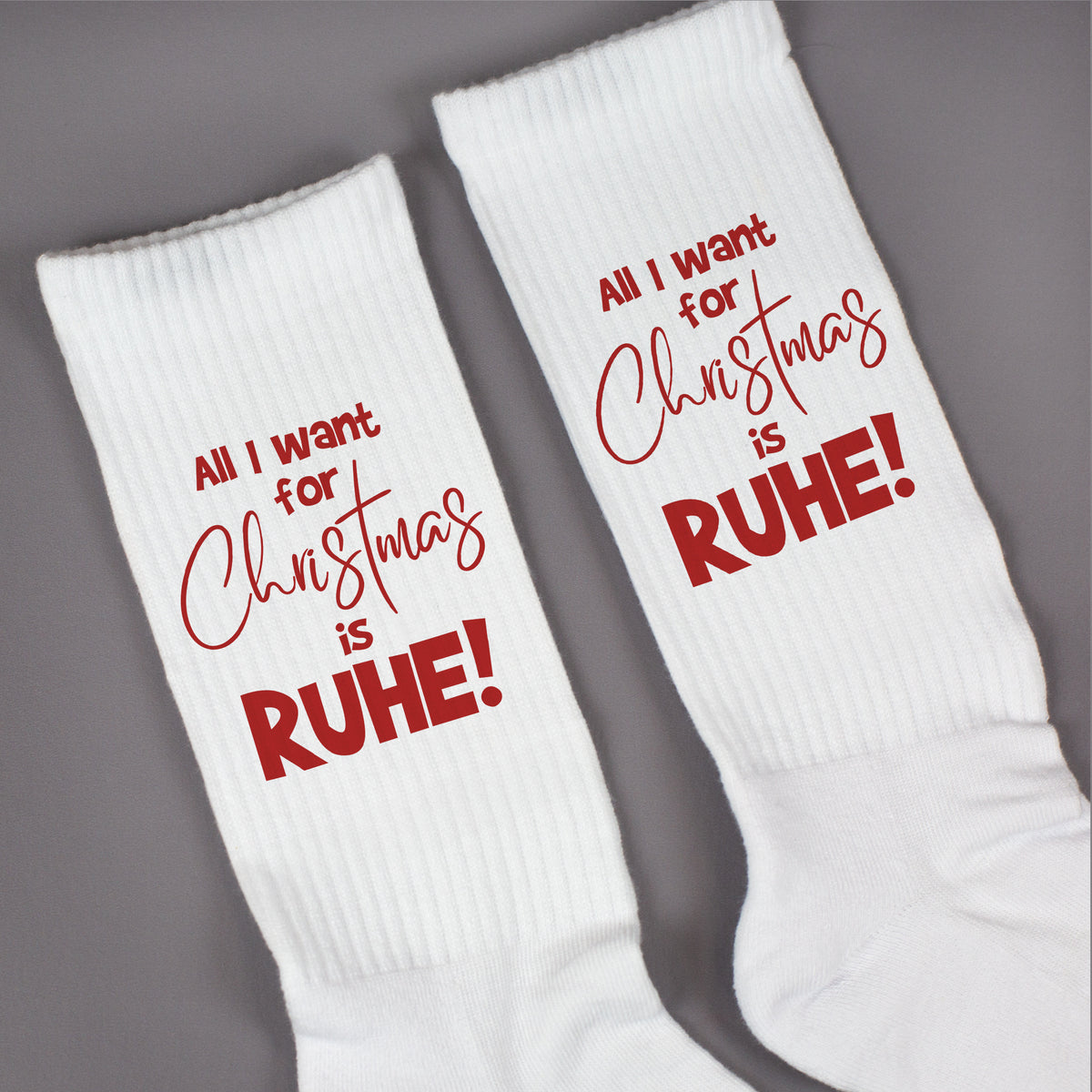 Paar weiße Tennissocken mit rotem Weihnachtsdruck ‚All I want for Christmas is RUHE!‘ flach nebeneinander liegend.