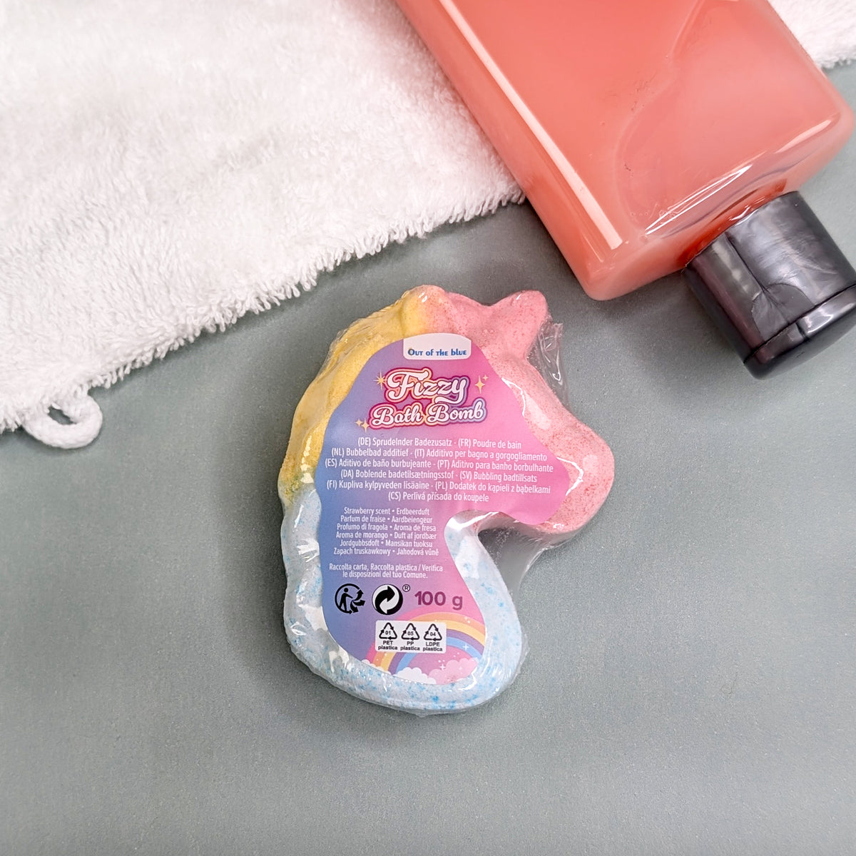 Verpackte Einhorn-Badekugel mit Etikett auf grauer Unterlage, daneben ein Handtuch und rosa Duschgel