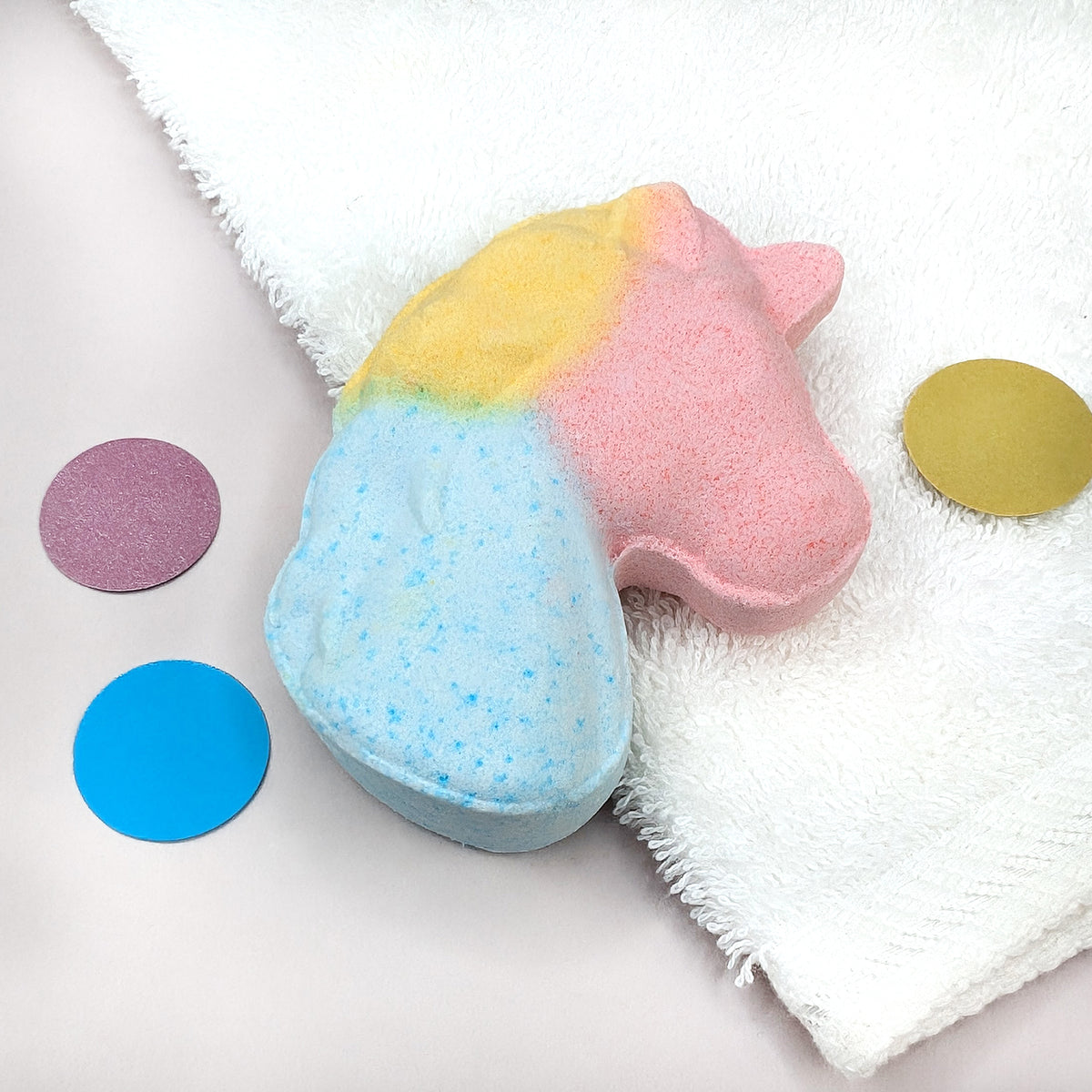 Bunte Einhorn-Badekugel in Rosa, Gelb und Blau auf weißem Handtuch mit bunten Dekopunkten