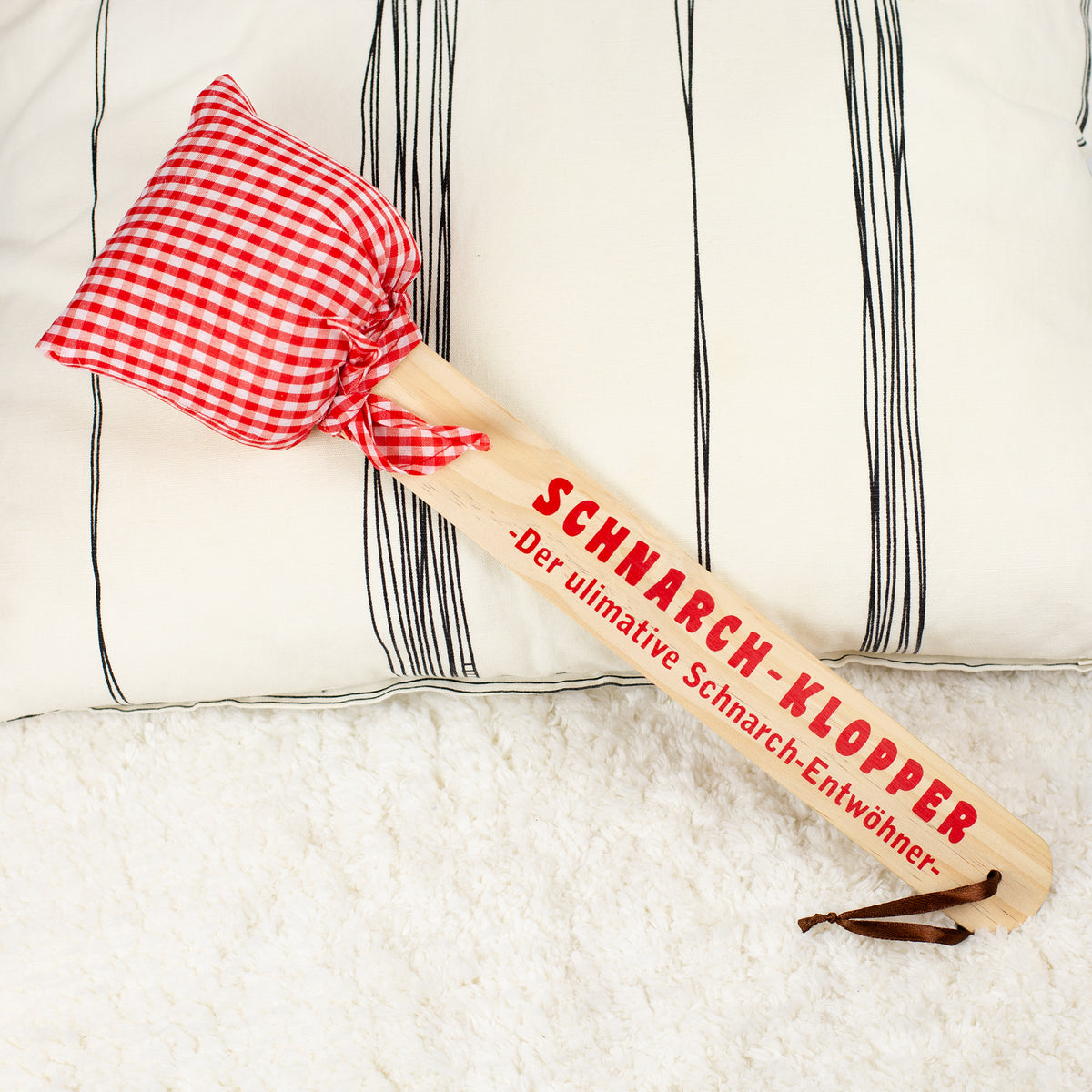 Schnarch-Klopper mit Holzgriff und rot-weiß kariertem Stoffkissen, Aufdruck „Der ultimative Schnarch-Entwöhner“, liegend auf hellem Untergrund