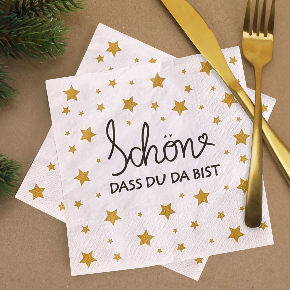 Weiße Servietten mit goldenem Sternenmuster und Schriftzug ‚Schön, dass du da bist‘, festlich mit goldfarbenem Besteck und Tannenzweigen dekoriert.