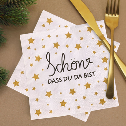 Weiße Servietten mit goldenem Sternenmuster und Schriftzug ‚Schön, dass du da bist‘, festlich mit goldfarbenem Besteck und Tannenzweigen dekoriert.