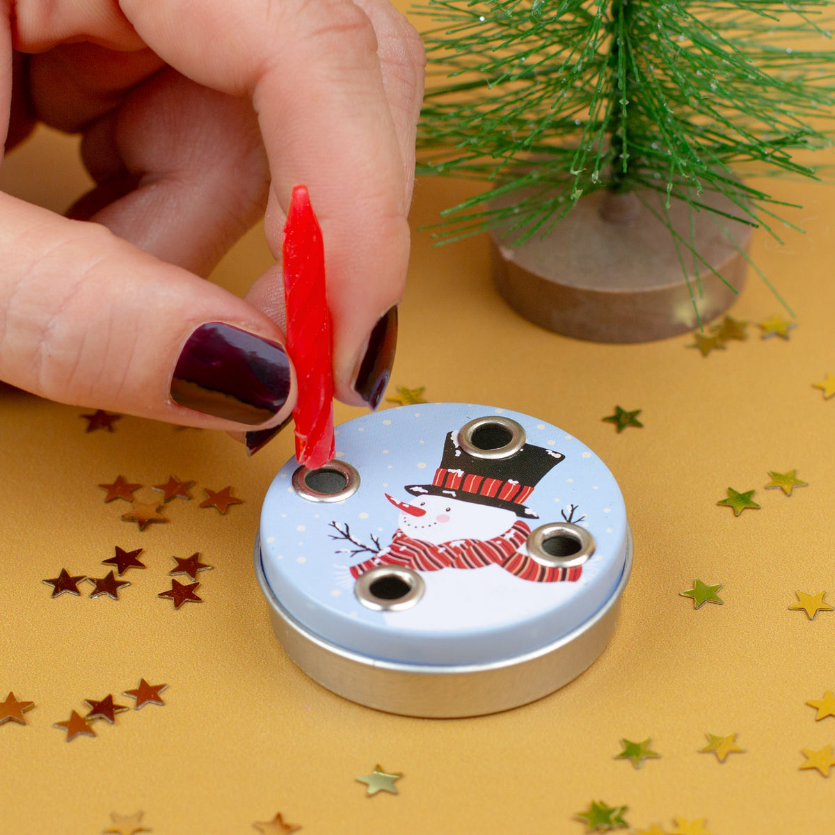 Nahaufnahme einer Hand, die eine rote Kerze in den Mini-Adventskranz mit Schneemannmotiv einsetzt, dekoriert mit kleinen Sternchen.