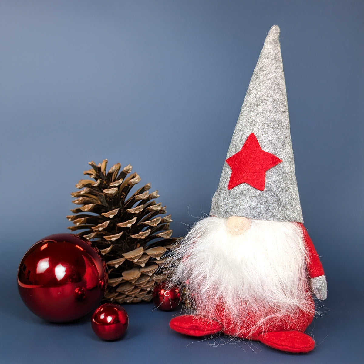 Weihnachtswichtel mit grauer Mütze und rotem Körper neben Tannenzapfen und Weihnachtskugeln