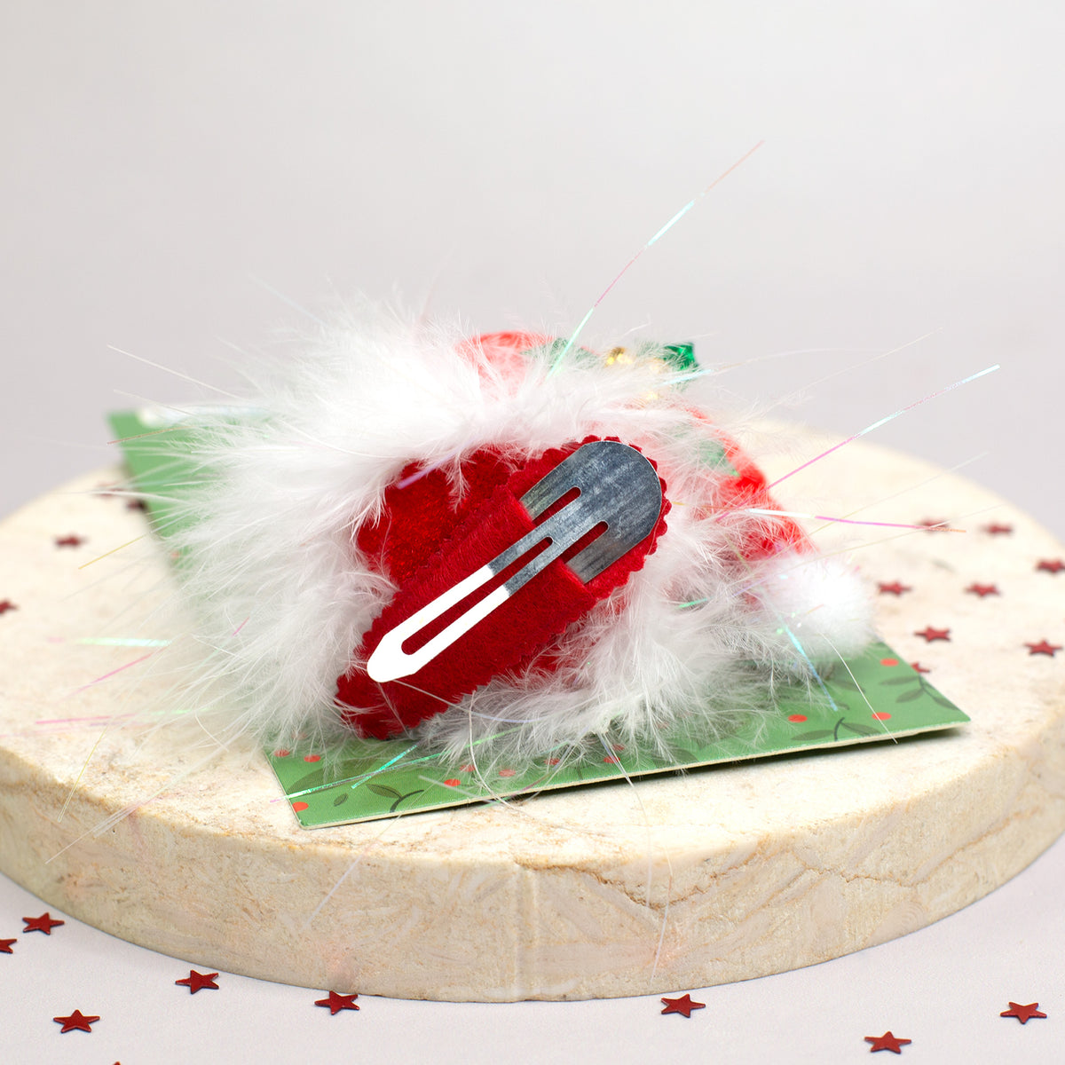 Weihnachtlicher Plüsch-Haarclip in Form einer roten Mütze mit Flaum und Metallclip, auf Deko-Untergrund mit Sternkonfetti.