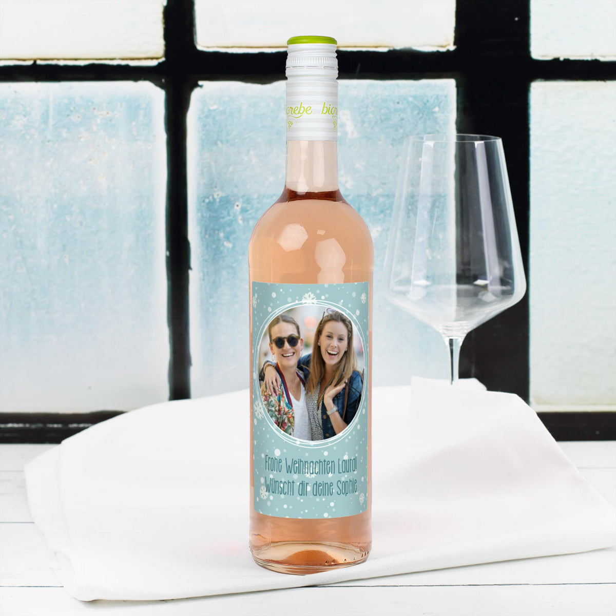 Personalisierte Roséweinflasche mit Schneeflocken-Etikett, eigenem Foto und Weihnachtsgruß – Szene vor Fenster