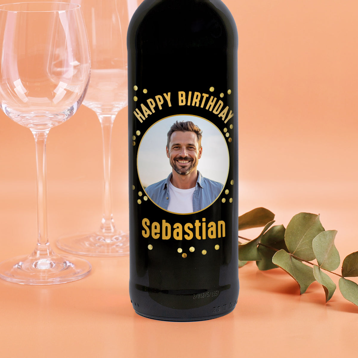 Weinflasche mit schwarzem Etikett „Happy Birthday Sebastian“ und Foto eines Mannes, dekoriert mit goldenen Punkten, vor Weingläsern.