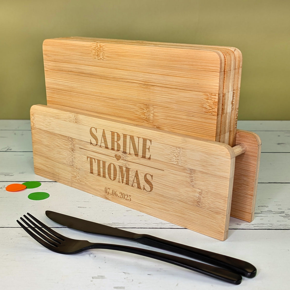 Bambusbrettchen-Set mit Gravur „Sabine & Thomas“ und Datum, daneben schwarzes Besteck auf hellem Tisch