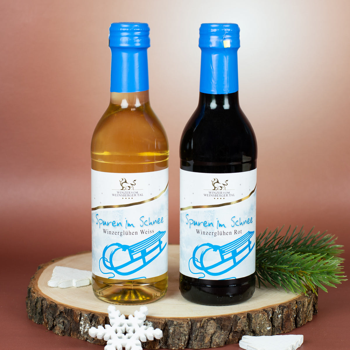 Flasche Winzerglühwein in Weiß und Flasche Winzerglühwein in Rot der Marke „Spuren im Schnee“, stehend auf einer Baumstammscheibe, dekoriert mit Tannenzweig und Schneeflocke vor warmem Hintergrund.