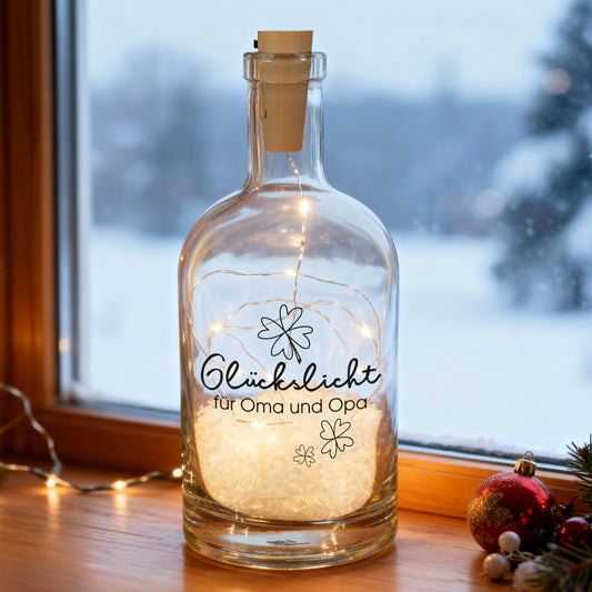 Beleuchtete Glückslicht-Karaffe mit Kleeblattmotiv und Wunschtext „für Oma und Opa“ auf einer Fensterbank, warmweiße LEDs im Inneren und winterlicher Schneelandschaft im Hintergrund.