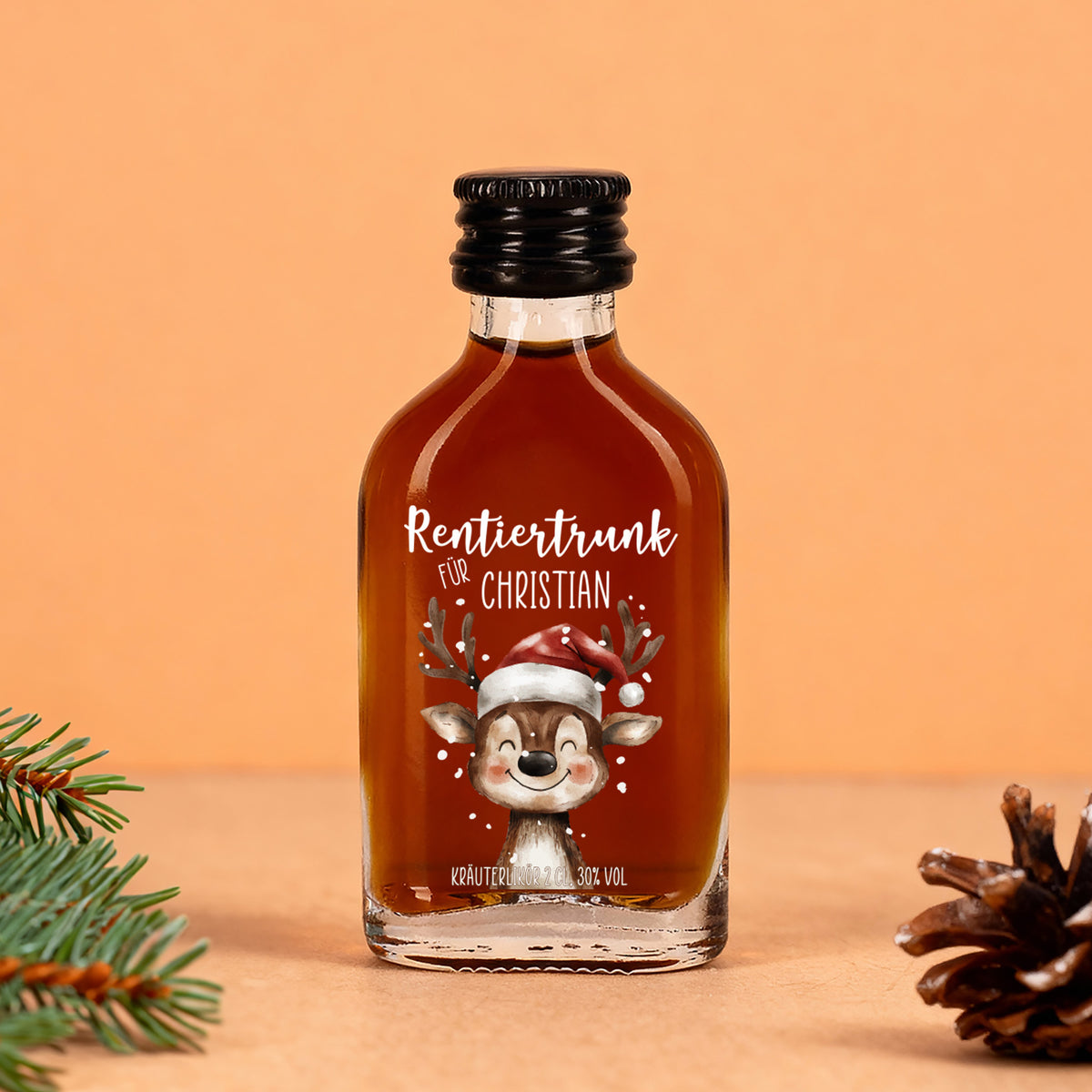 Mini-Flasche mit braunem Kräuterlikör, Aufdruck „Rentiertrunk für Christian“, Rentier mit Mütze, arrangiert mit Tannenzweig und Zapfen auf warmem Hintergrund.
