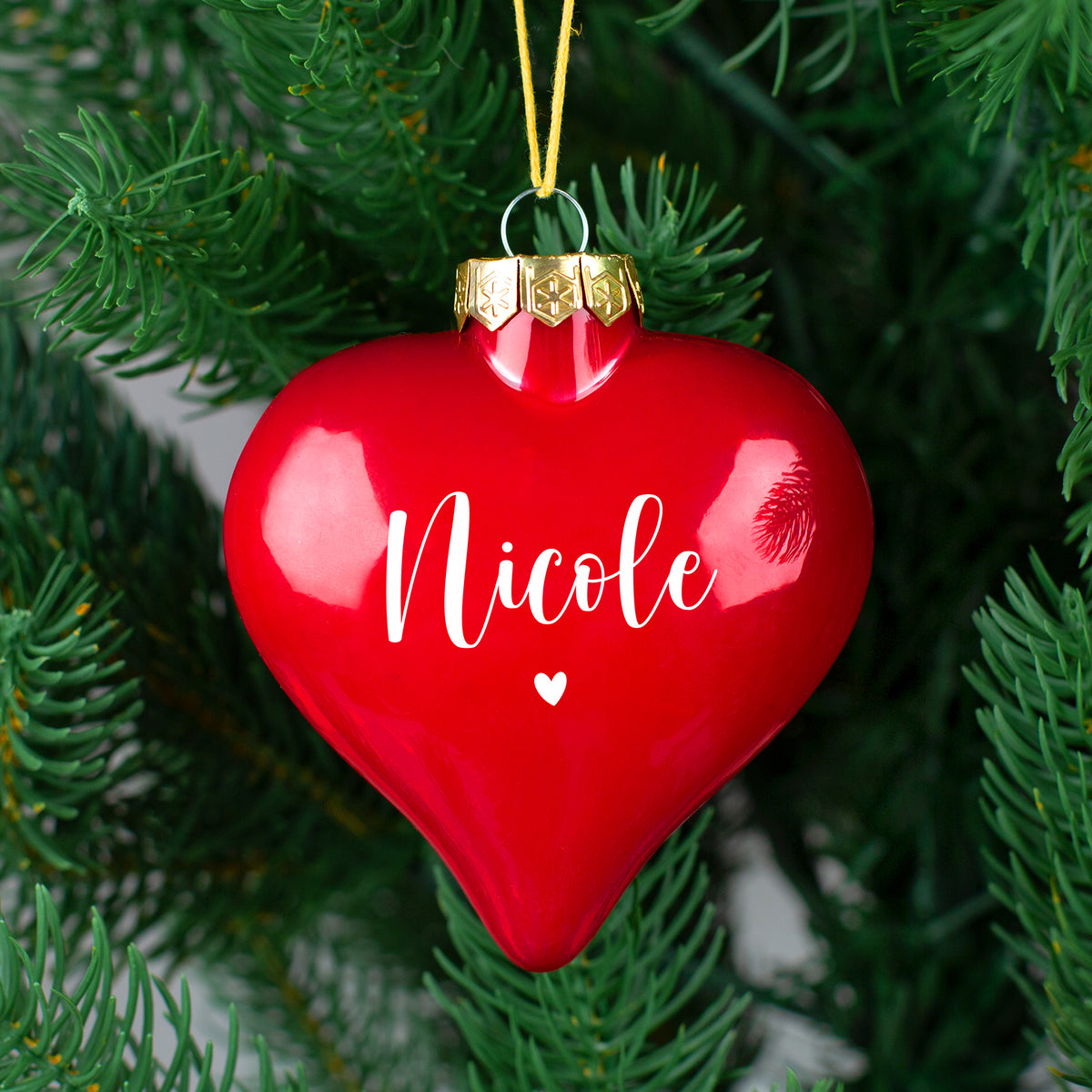 Rote, glänzende Herz-Christbaumkugel mit weißem Namen „Nicole“ und kleinem Herz, goldene Metallöse, am Tannenzweig hängend.
