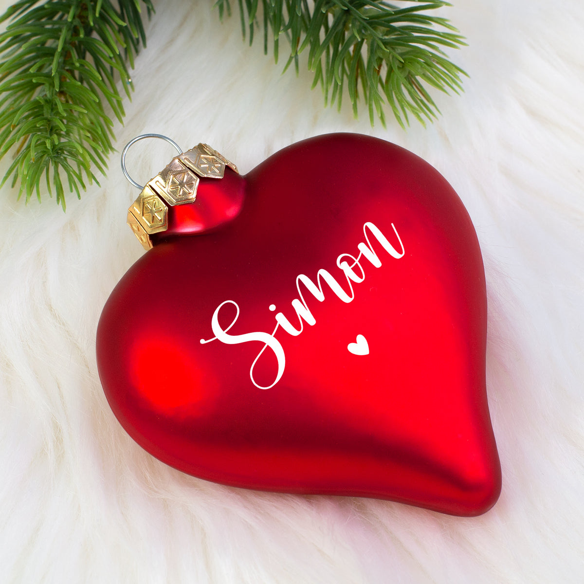 Matte rote Herz-Weihnachtskugel mit Name „Simon“ liegend auf weißem Fell, goldene Aufhängekappe sichtbar.