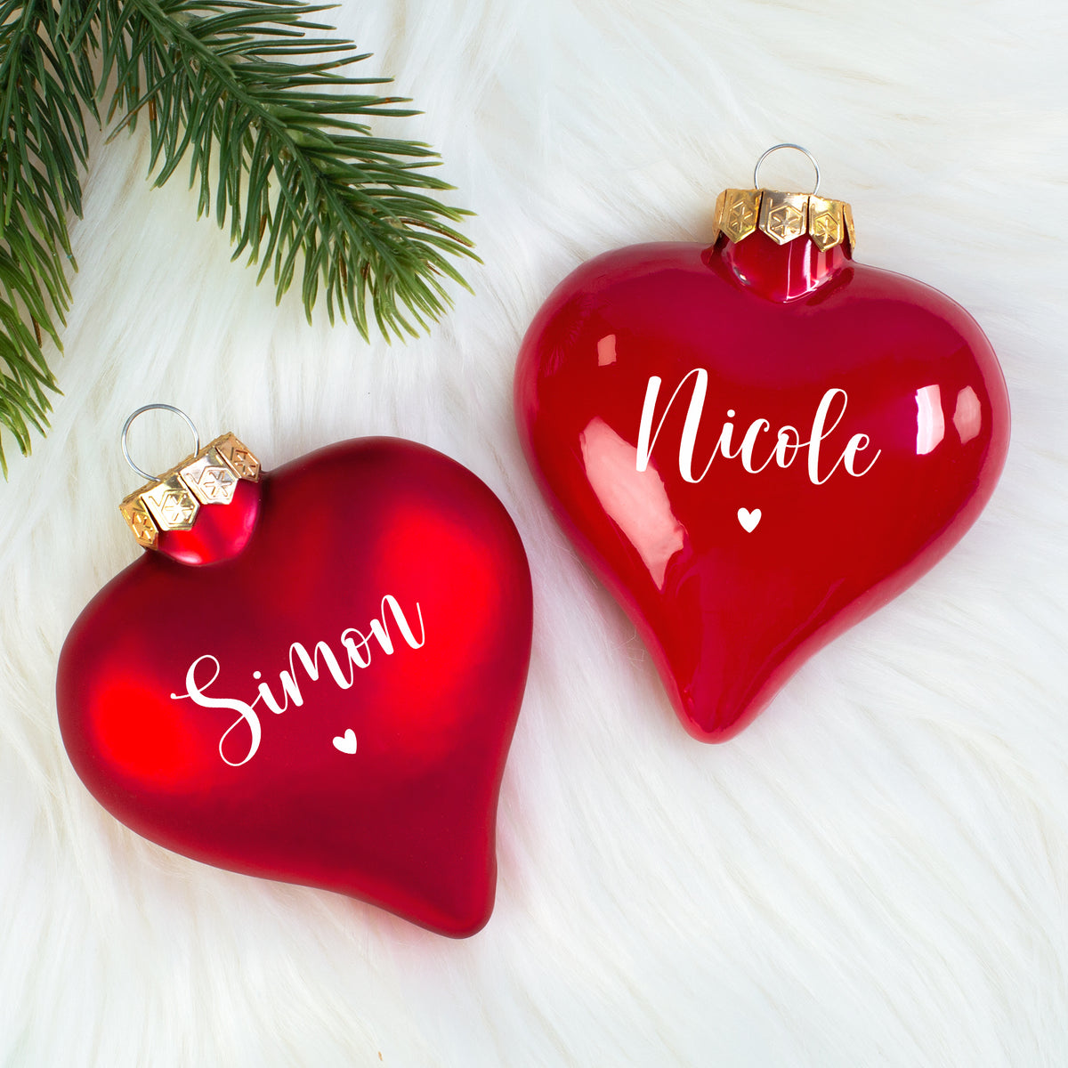 Zwei personalisierte Herz-Christbaumkugeln (matt „Simon“, glänzend „Nicole“) nebeneinander auf weißem Fell mit Tannenzweig.