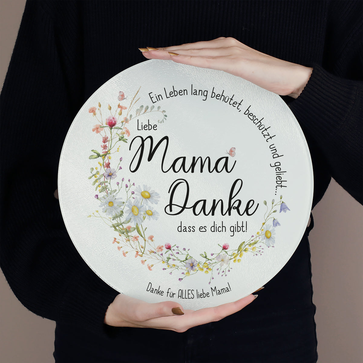 Eine Person hält das Glasbrettchen mit beiden Händen vor den Körper. Das transparente Material mit strukturierter Oberfläche ist gut sichtbar. Die Botschaft „Mama, Danke, dass es dich gibt!“ ist von einem detailreichen Blumenkranz und dem Schriftzug „Ein Leben lang behütet, beschützt und geliebt…“ umrahmt. Der Wunschtext unten lautet „Danke für ALLES liebe Mama!“.