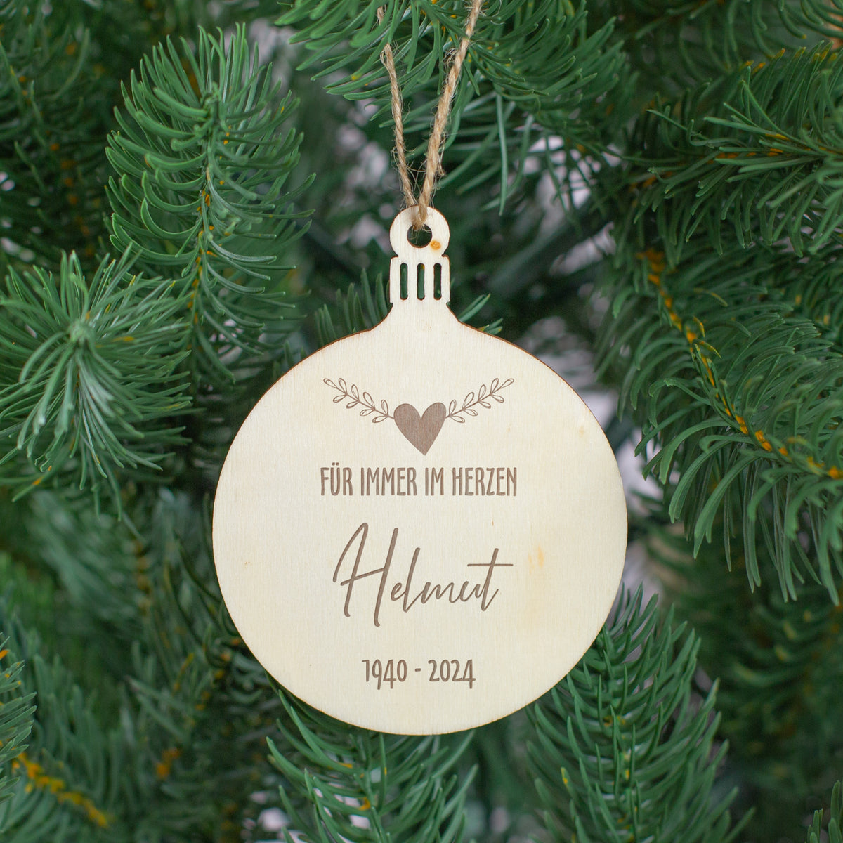 Erinnerungsanhänger aus Holz mit Gravur „Für immer im Herzen“, Name Helmut und Jahreszahlen 1940–2024, hängend am Weihnachtsbaum zwischen grünen Zweigen.