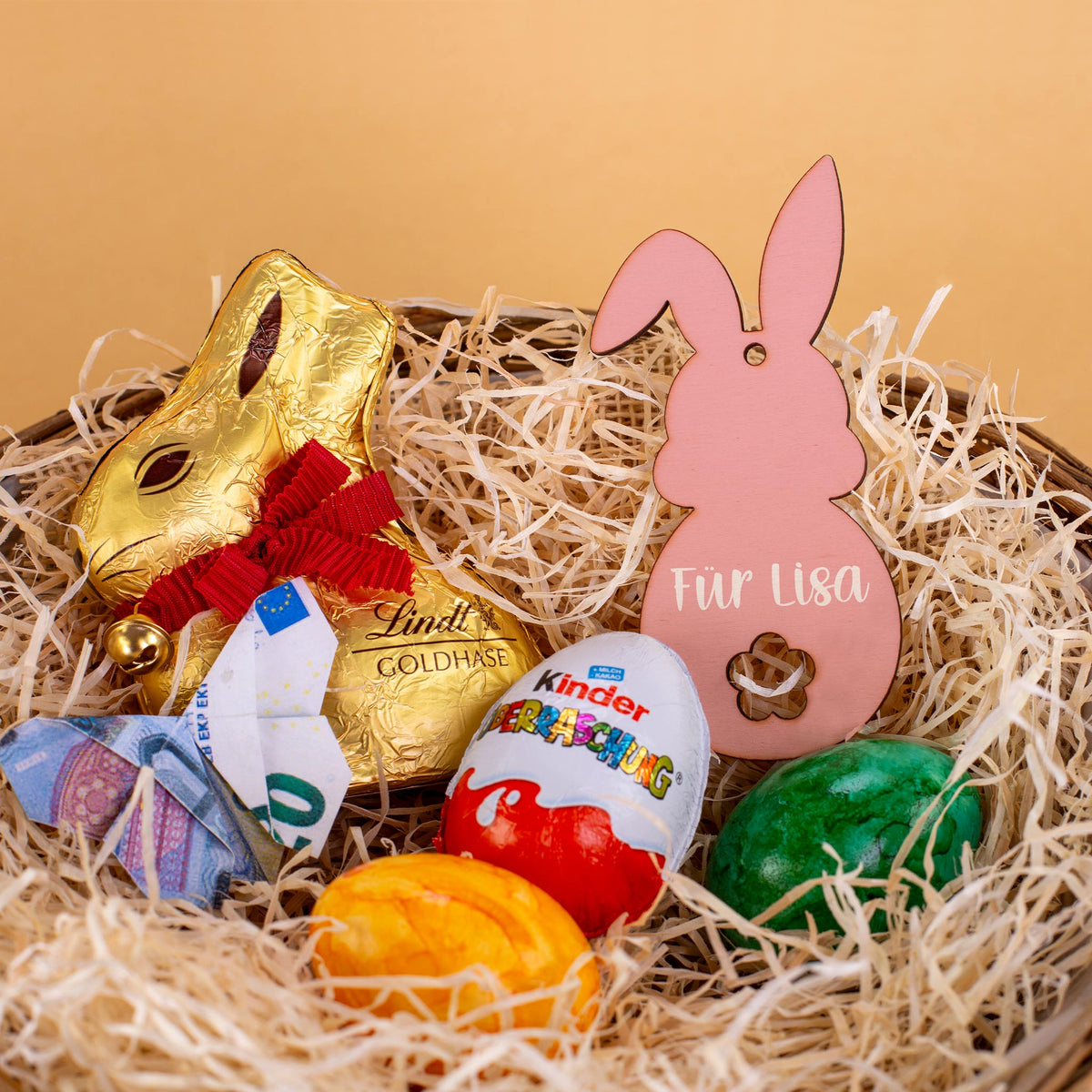 Personalisierter Holzanhänger in Rosa als Dekoration in einem Osternest mit Schokolade und Geldschein, bedruckt mit Wunschtext