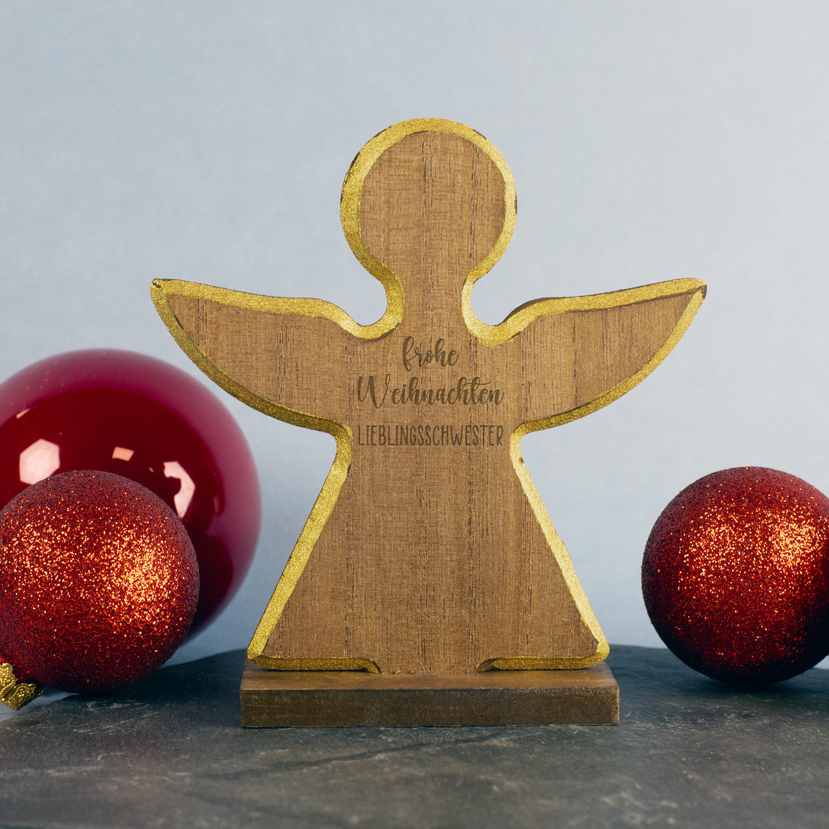Holzengel mit Gravur Frohe Weihnachten Lieblingsschwester, goldene Kante, rote Weihnachtskugeln im Hintergrund