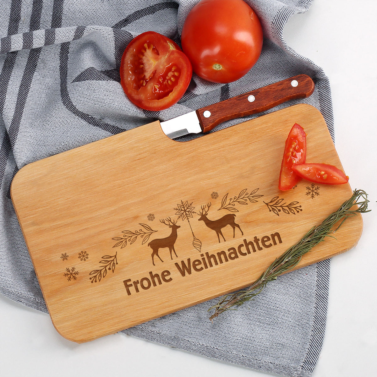 Frühstücksbrettchen aus Holz mit Gravur „Frohe Weihnachten“, dekoriert mit Tomaten und Rosmarin auf grauem Tuch