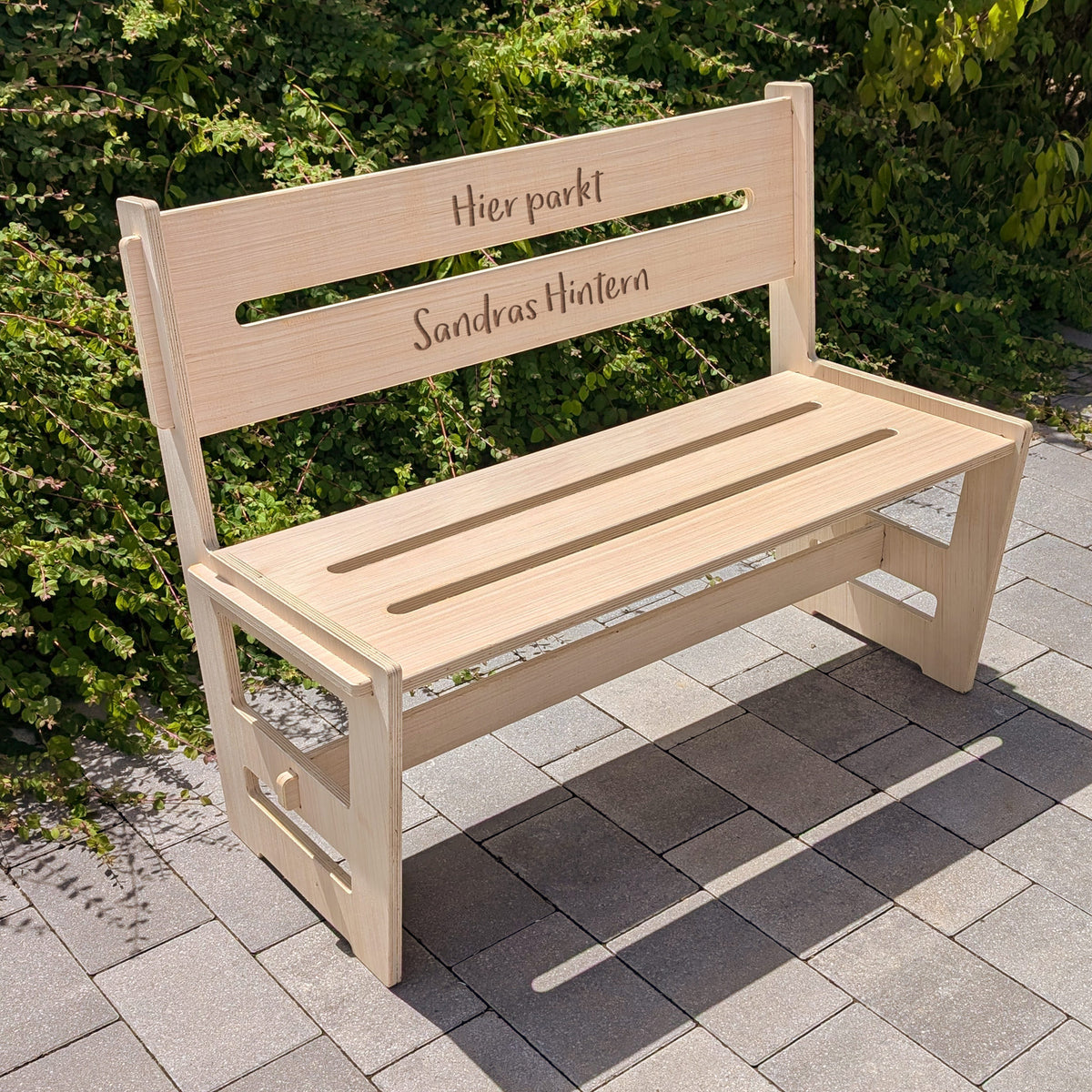 Personalisierte Holzbank mit Gravur „Hier parkt Sandras Hintern“, im Garten auf gepflastertem Untergrund fotografiert