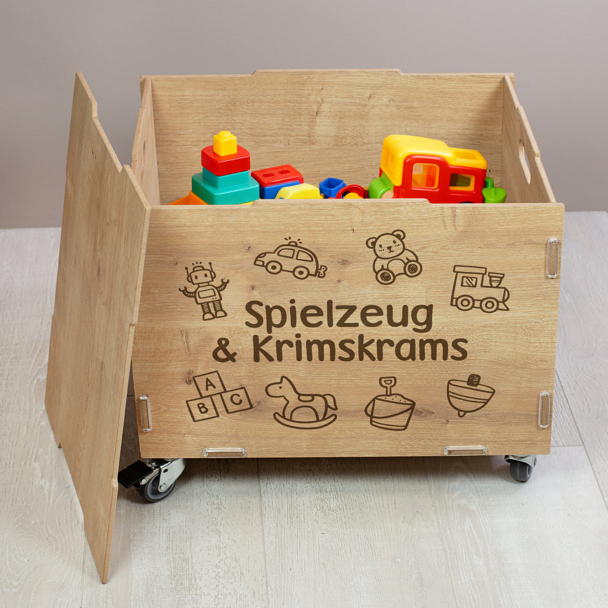 Geöffnete personalisierte Rollkiste aus Holz mit Spielzeug im Inneren, Vorderseite mit graviertem Wunschtext „Spielzeug & Krimskrams“ und kindlichen Symbolen