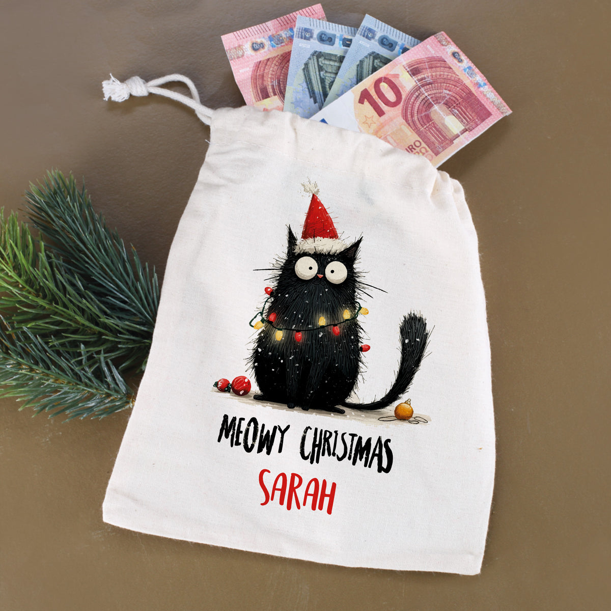 Stoffbeutel mit Katzenmotiv „Meowy Christmas – Sarah“, befüllt mit Geldscheinen und Tannenzweig daneben