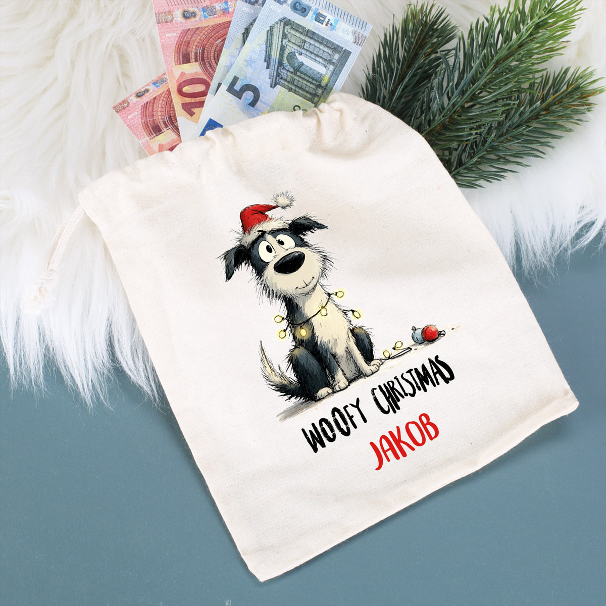 Stoffbeutel mit Hundemotiv „Woofy Christmas – Jakob“, befüllt mit Geldscheinen, auf weißem Fell mit Tannenzweig