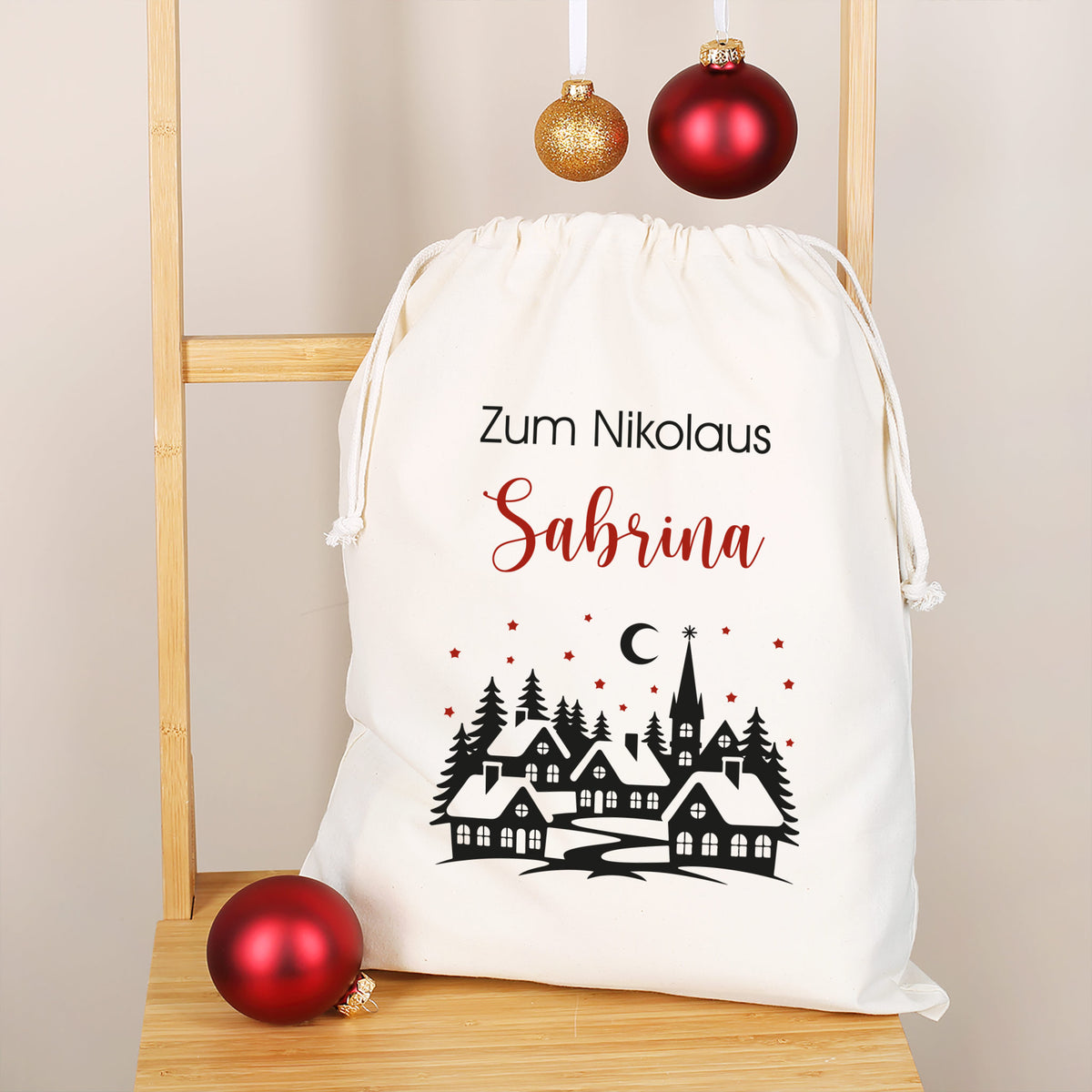 Großer Nikolausbeutel mit Wunschtext „Zum Nikolaus Sabrina“, verschneite Dorfkulisse mit Tannen und Sternenhimmel, goldrote Kugeln als Deko.