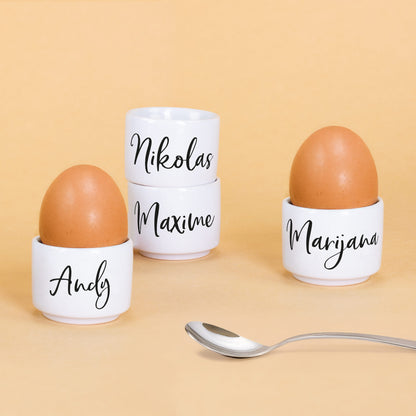 Vier weiße Keramik-Eierbecher mit Eiern und Namen „Andy“, „Marijana“, „Nikolas“ und „Maxime“ aufgelistet auf gelbem Hintergrund