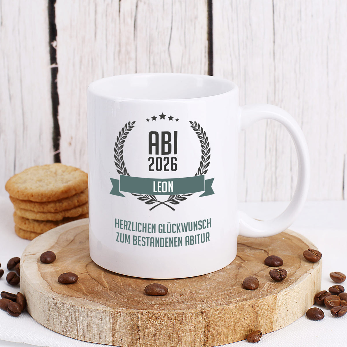 Glückwunsch-Tasse zum Abitur mit Name und Wunschtext dekoriert auf Baumscheibe mit Keksen und Kaffeebohnen