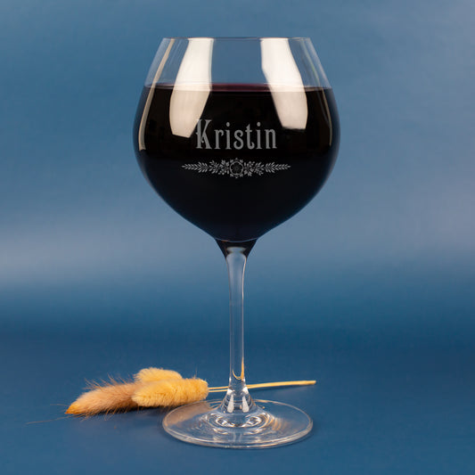 Personalisiertes Leonardo Weinglas mit Gravur des Namens Kristin und floralem Ornament – edles Burgunderglas mit Rotwein, vor blauem Hintergrund mit zarten Trockenhalmen.