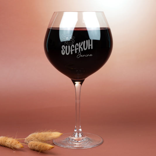 Burgunderglas von Leonardo mit Gravur „Suffkuh“ und Name Janina, gefüllt mit Rotwein – humorvolles Weinglas mit Kuh-Motiv, fotografiert auf braunem Hintergrund mit Trockenblumen.