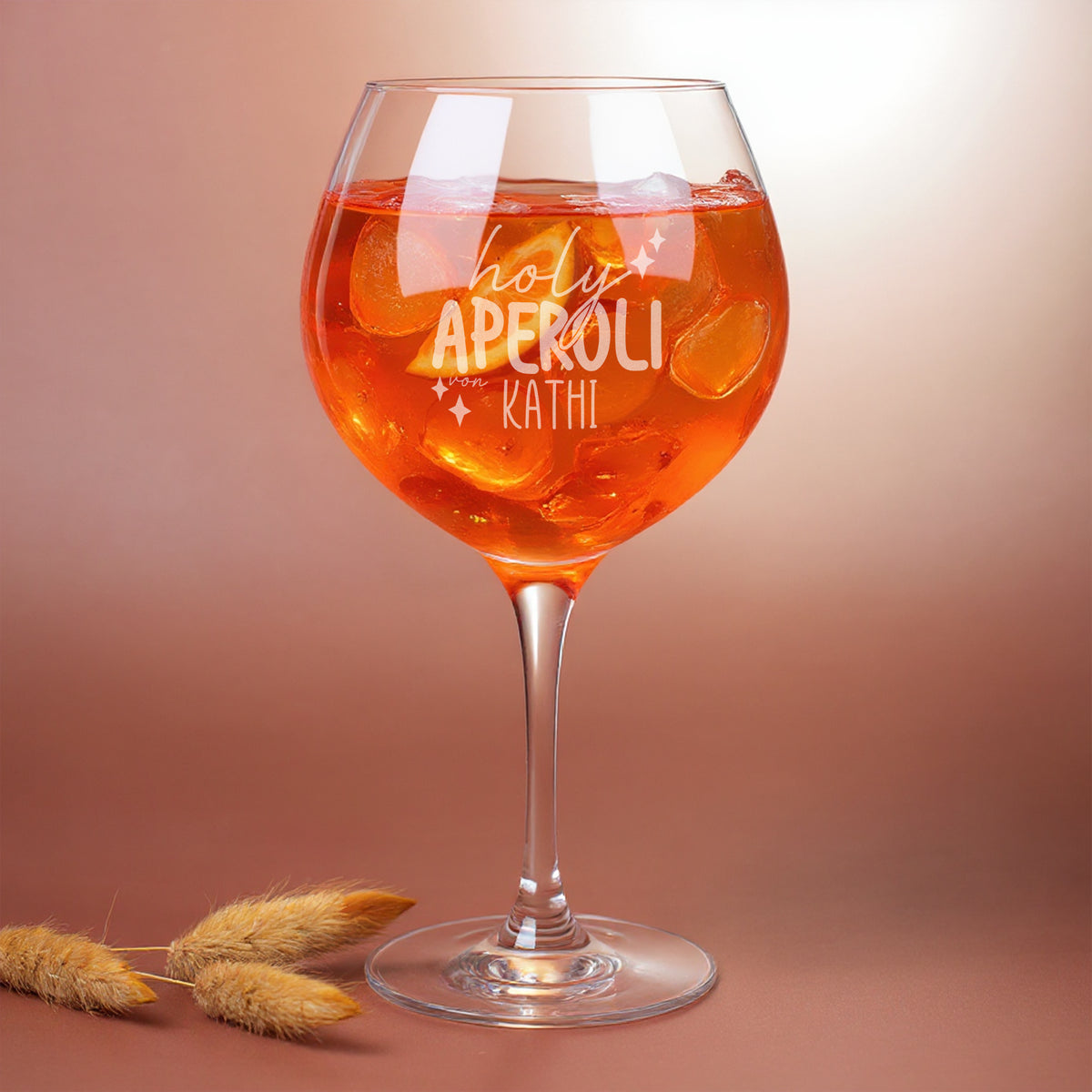 Leonardo Weinglas mit Gravur „holy aperoli von Kathi“, gefüllt mit Eiswürfeln und Aperitifgetränk – humorvolles, personalisiertes Glas für Sommerdrinks vor warmem Hintergrund.