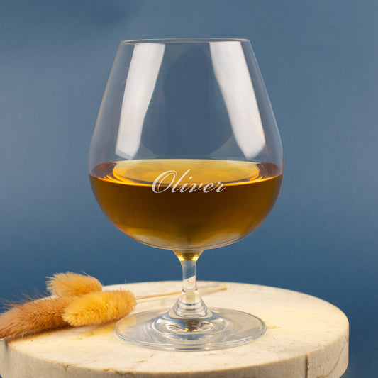 Leonardo Cognacglas mit Gravur „Oliver“ – auf hellem Holzuntergrund mit Trockenblumen, blauer Hintergrund.