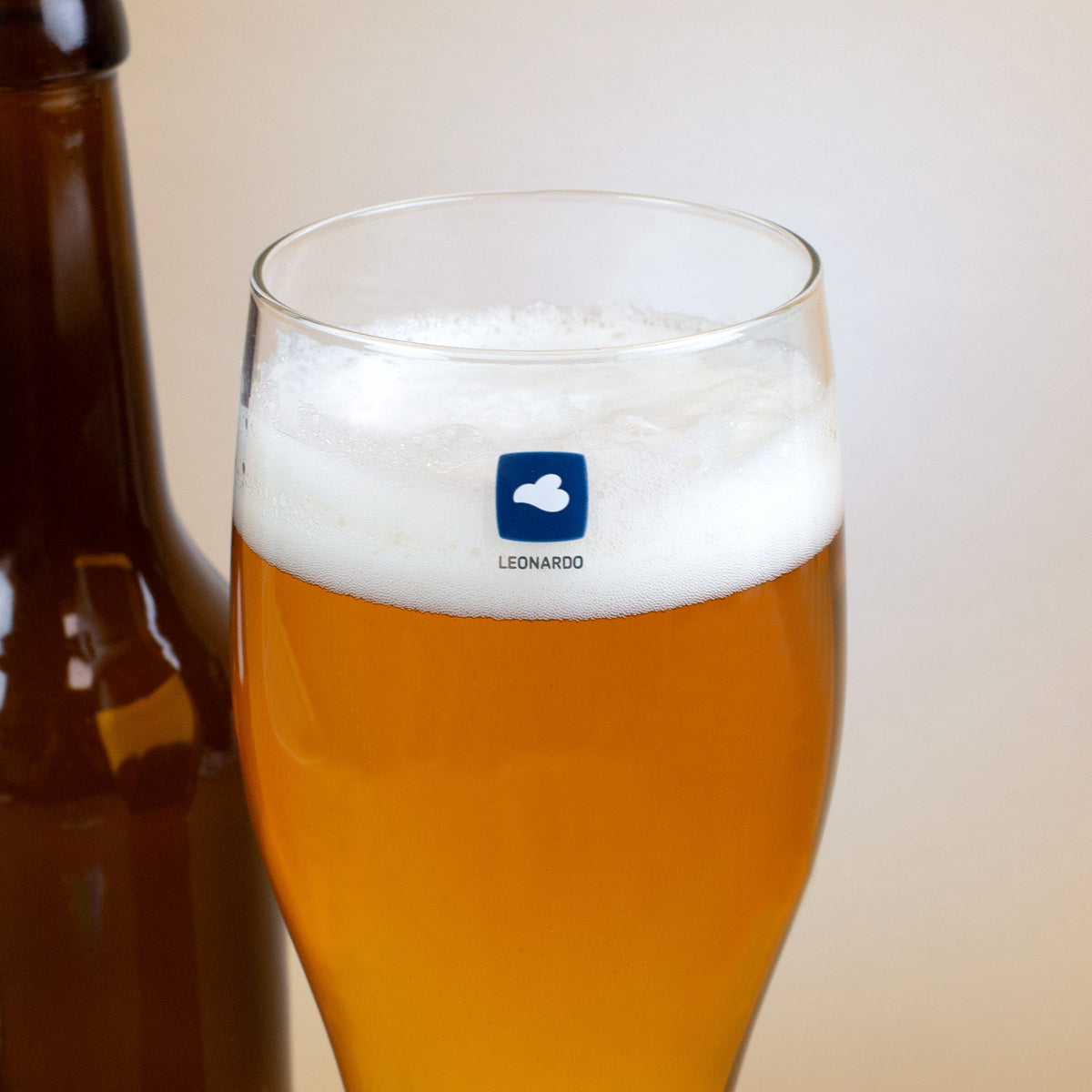 Detailaufnahme eines Leonardo Bierglases mit blauem Logo und Schaumkrone, im Hintergrund eine Bierflasche