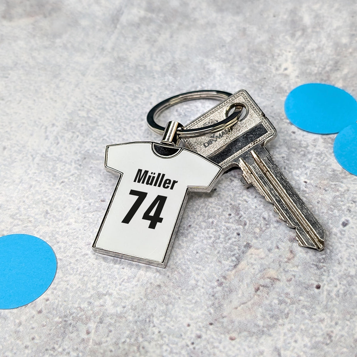 Weißer Trikot-Schlüsselanhänger mit Namen „Müller“ und Zahl „74“ an einem Schlüsselring auf hellem Untergrund