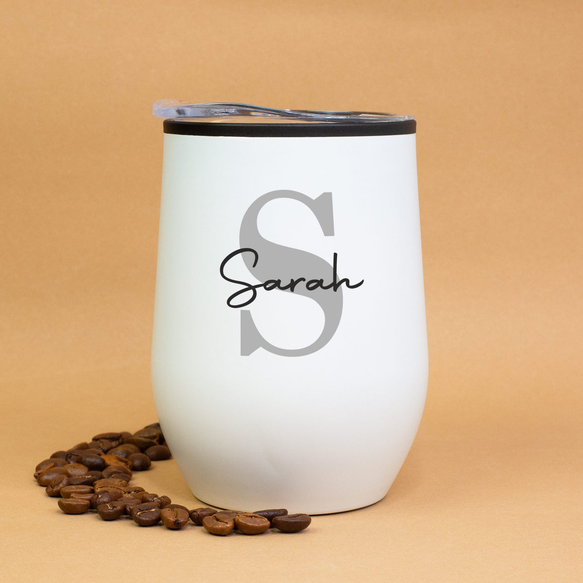 Weißer Thermobecher mit Initiale S und Name Sarah, auf braunem Hintergrund mit Kaffeebohnen dekoriert