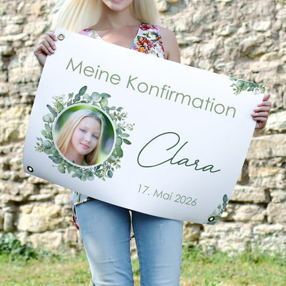 Mädchen hält ein Banner mit ihrem Foto im Eukalyptuskranz, beschriftet mit „Meine Konfirmation – Clara – 17. Mai 2026“. Outdoor-Aufnahme vor Natursteinmauer, mit Metallösen in den Ecken.