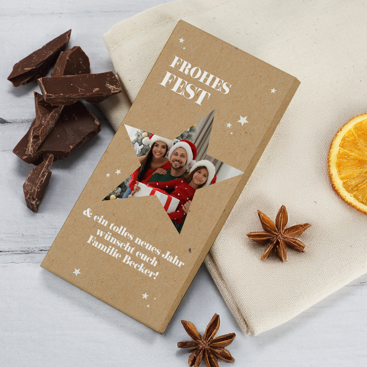 Foto-Schokoladentafel mit Sternmotiv und weihnachtlichem Design. Aufdruck „Frohes Fest“ über einem Foto einer Familie mit Weihnachtsmützen, integriert im Stern. Darunter persönlicher Grußtext. Daneben liegen Schokoladenstücke, getrocknete Orange und Sternanis auf hellem Tuch.