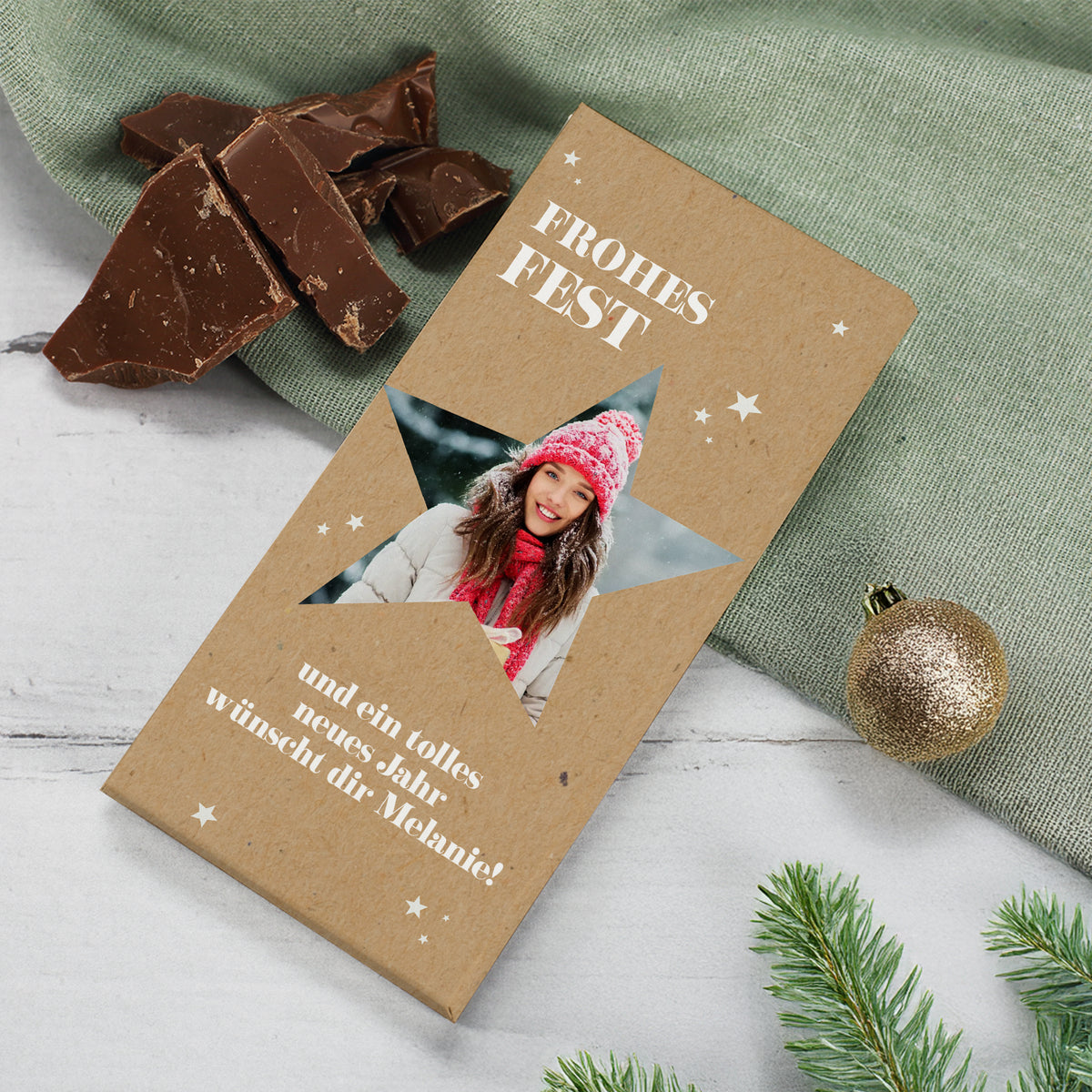 Weihnachtsschokolade mit Foto im Stern und individuellem Text. Verpackung in Kraftpapieroptik mit weißen Sternen. Im Ausschnitt ein winterliches Porträt einer Frau mit Mütze und Schal. Dekoriert mit grüner Stoffunterlage, Schokoladenstücken, goldener Kugel und Tannenzweig.